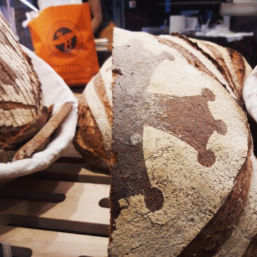 A close-up of rustic round loaves with dark, crusty tops and a crown-shaped flour pattern, displayed on a wooden shelf at the Victor Hugo Market bakery. An orange bag is visible in the background, adding to the lively market tour ambiance.