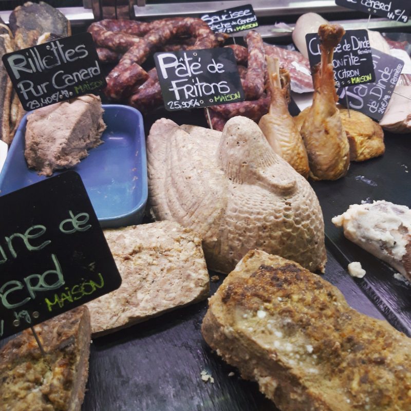 A display of French deli meats and pâtés, including a duck-shaped pâté, terrines, and sausages, awaits at Victor Hugo Market in Toulouse. Handwritten chalkboard labels in French describe the different meats in the showcase.