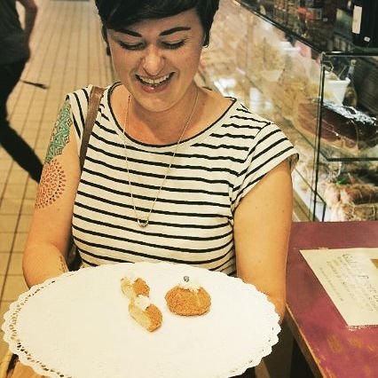 A woman in a striped shirt smiles while holding a large tray with just a few small pastries at Victor Hugo Market. She has short dark hair and a colorful tattoo on her upper arm, capturing the charm of this bustling market tour.