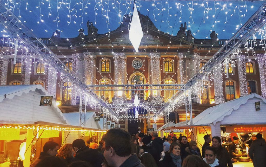 A festive outdoor market at night, part of November events, with crowds walking between white stalls illuminated by strings of lights. In the background, a grand building with columns and a clock completes the cheerful scene.