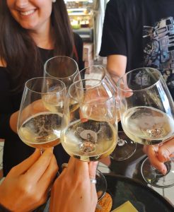 Five people clink glasses of white wine together in a toast during a casual, festive gathering. One woman smiles as cheese and crackers sit on the table below—perhaps celebrating November events or sharing a news roundup with friends.