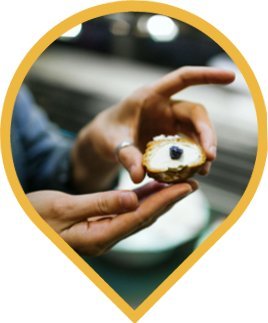 A person holds a small piece of bread or cracker topped with cream and a single blueberry, with a blurred background. A person holds a small piece of bread or cracker topped with cream and a single blueberry, with a blurred background.
