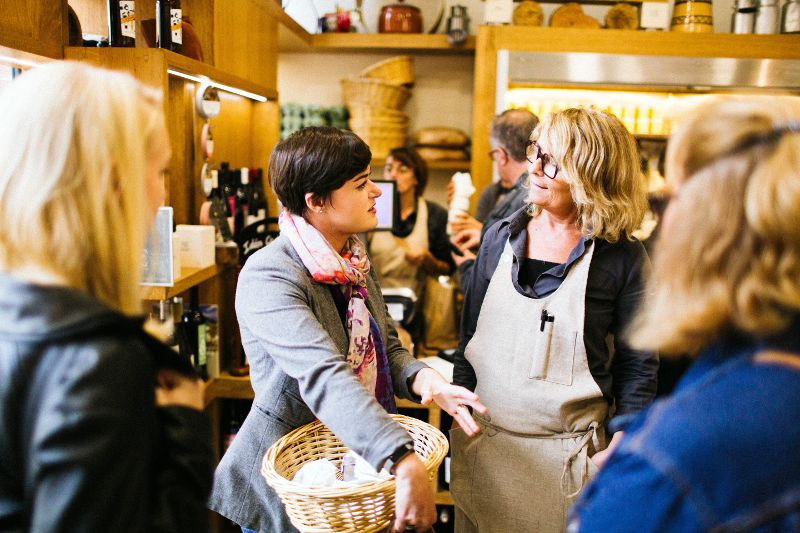 Two women, one holding a wicker basket and the other wearing an apron, talk inside a busy shop with shelves of products and several people in the background.
