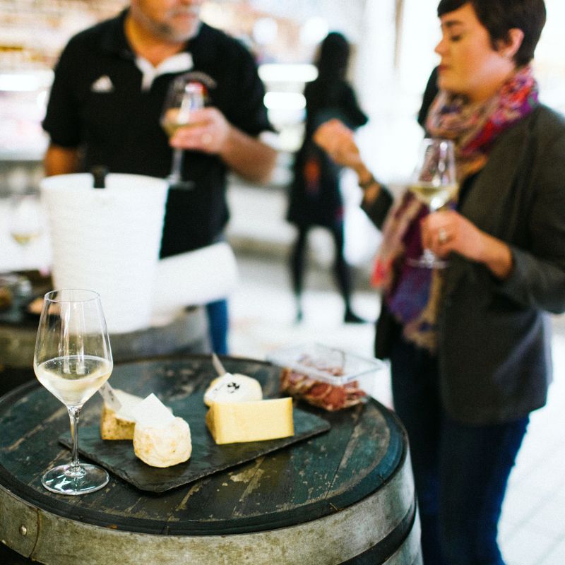 Two people stand by a barrel table with a platter of cheese and charcuterie, each holding a glass of white wine. The setting appears casual and social, with blurred people and shelves in the background.
