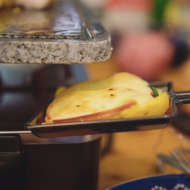 A close-up of a spatula holding a slice of melted raclette cheese over meat and vegetables, being heated under a raclette grill. The background is blurred with kitchen items.