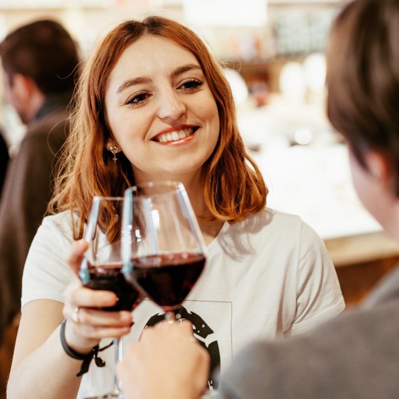 A smiling woman with red hair clinks glasses of red wine with another person in a lively indoor setting, capturing the spirit of Taste of Toulouse.