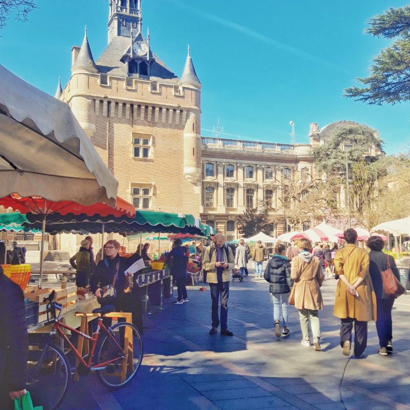 People stroll and shop at an outdoor market in Toulouse, browsing food events under colorful tents in a sunny plaza. A historic brick building with a tall clock tower stands nearby, while trees and blue sky frame the lively scene.