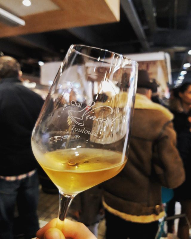 A hand holds a wine glass with a small amount of golden liquid inside, labeled "Chai l’Inconnu Toulouse," as people mingle at a busy indoor market—an inviting scene for those looking to buy wine in Toulouse.
