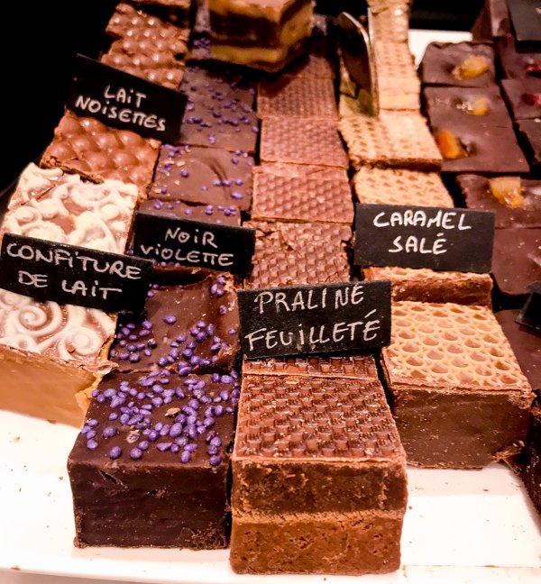 Assorted chocolate bars with various textures and toppings, each labeled with handwritten signs in French, showcase the rich food culture of Toulouse, featuring flavors like praline feuilleté, caramel salé, lait noisettes, and confiture de lait.