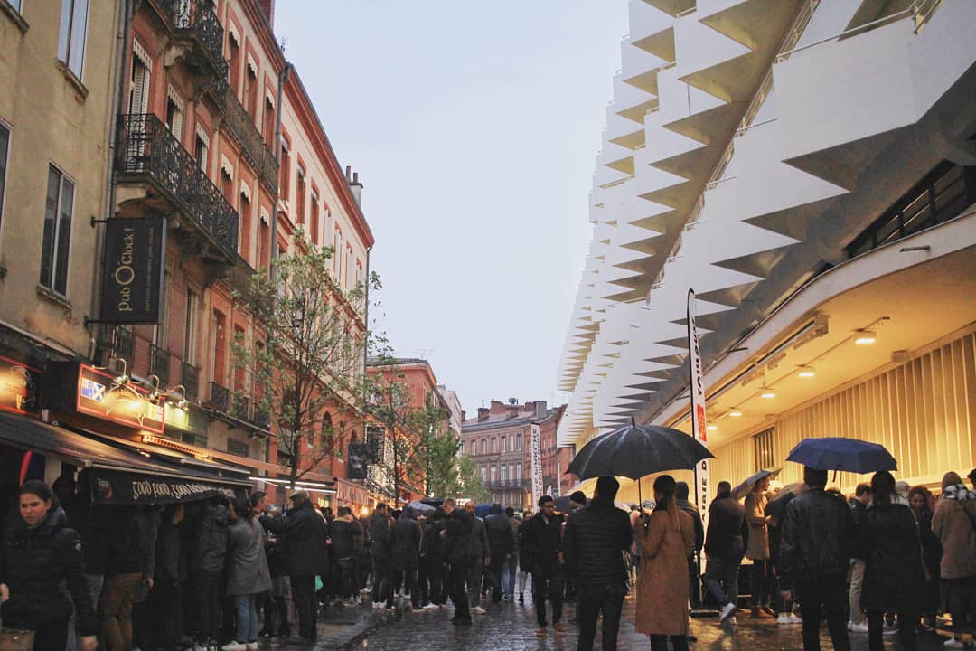 A crowd with umbrellas walks on a wet city street lined with cafes and shops; modern white architecture contrasts with older red-brick buildings on a rainy day—perfect for reading our May Newsletter or catching up on local events.