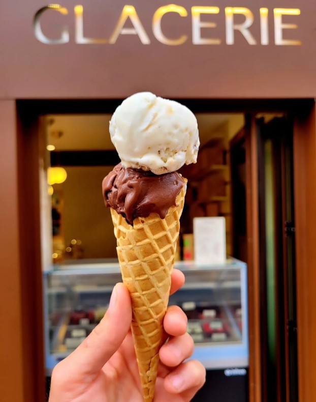 A hand holds a waffle cone with two scoops of ice cream, chocolate and vanilla, in front of a shop with a sign that reads “GLACERIE,” capturing the essence of enjoying classic ice cream in Toulouse.