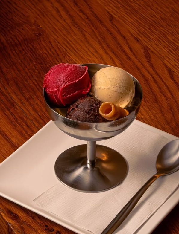 A metal dessert cup with three scoops of ice cream—red, beige, and dark brown, including a classic ice cream Toulouse—garnished with rolled wafer cookies, sits on a napkin-covered plate next to a spoon on a wooden table.