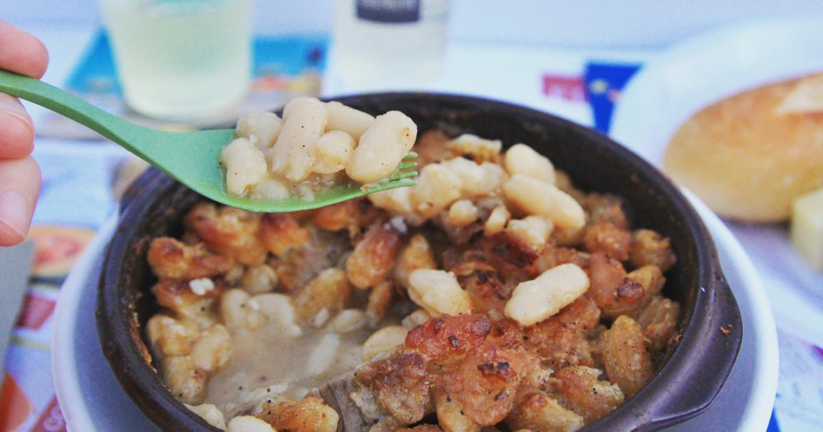 A close-up of a bowl of baked white beans with crispy browned topping, with a green spoon lifting a portion—perfect inspiration for our Cassoulet Giveaway contest. A bread roll and drink are blurry in the background.