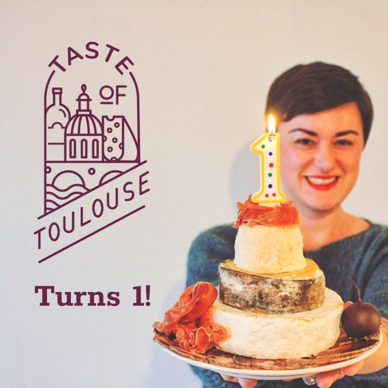 A smiling woman holds a cheese wheel “cake” with a lit number 1 candle. Beside her, the Taste of Toulouse logo and "Turns 1!" text pop against a light background—celebrating this milestone in local Toulouse news.