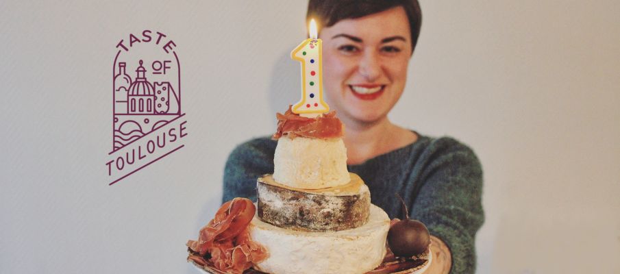 A smiling person holds a cheese wheel “cake” stacked in three tiers, topped with a number 1 candle and garnished with cured meat—perfect for any wine lover's party. The "Taste of Toulouse" logo appears on the left side of the image.