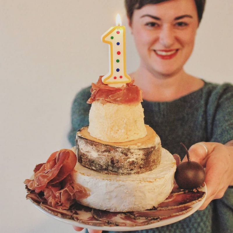 A smiling woman holds a plate with a savory cheese wheel "cake" topped with cured meat and a lit number 1 candle, perfect for a unique Taste of Toulouse 1st Birthday celebration. The focus is on the cake, with the woman slightly blurred in the background.