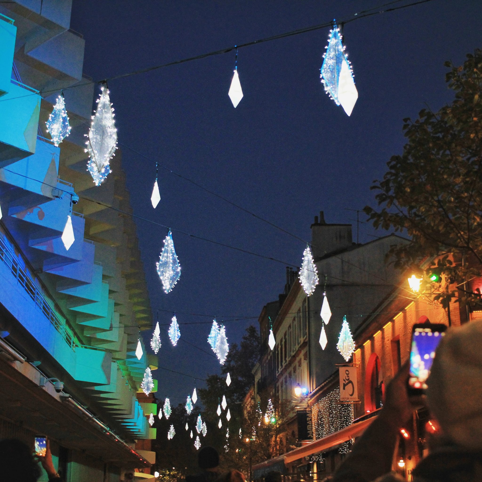 Hanging crystal-shaped lights glow above a city street at night. People below, some taking photos, enjoy the festive decoration—perfect for December food events. Buildings on both sides are illuminated, with trees visible on the right.