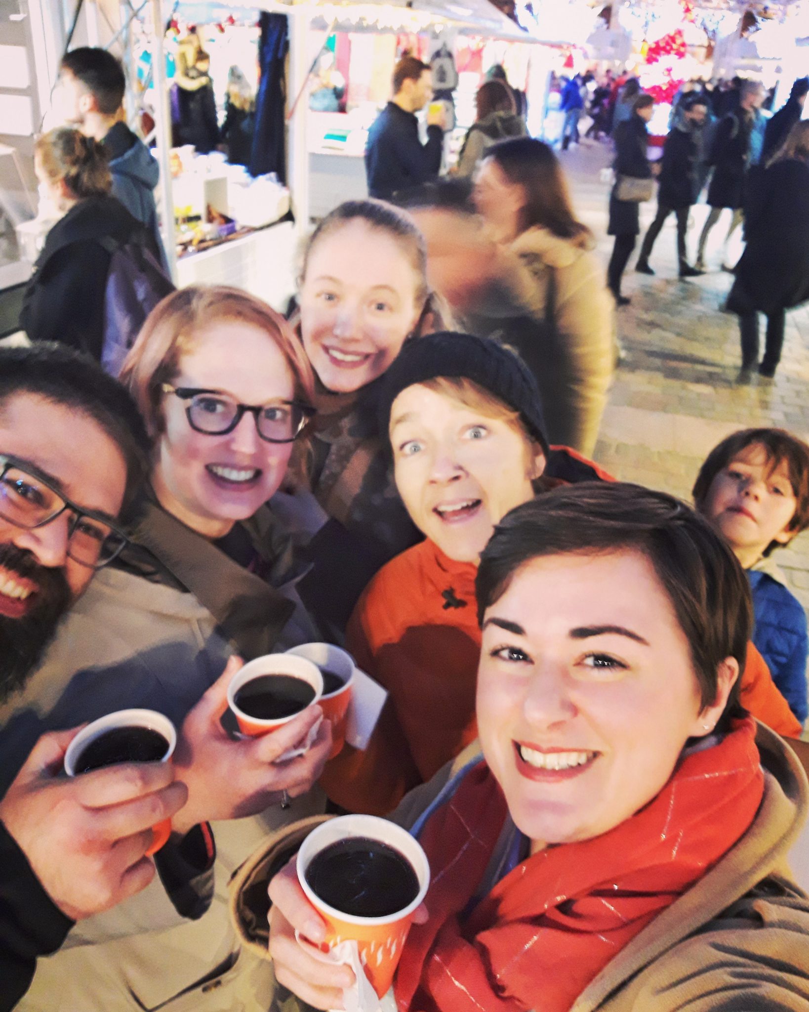 A group of five smiling adults hold hot drinks and pose for a selfie at a festive Christmas Market meetup, with bright lights and people in the background. A child stands nearby, looking at the camera.