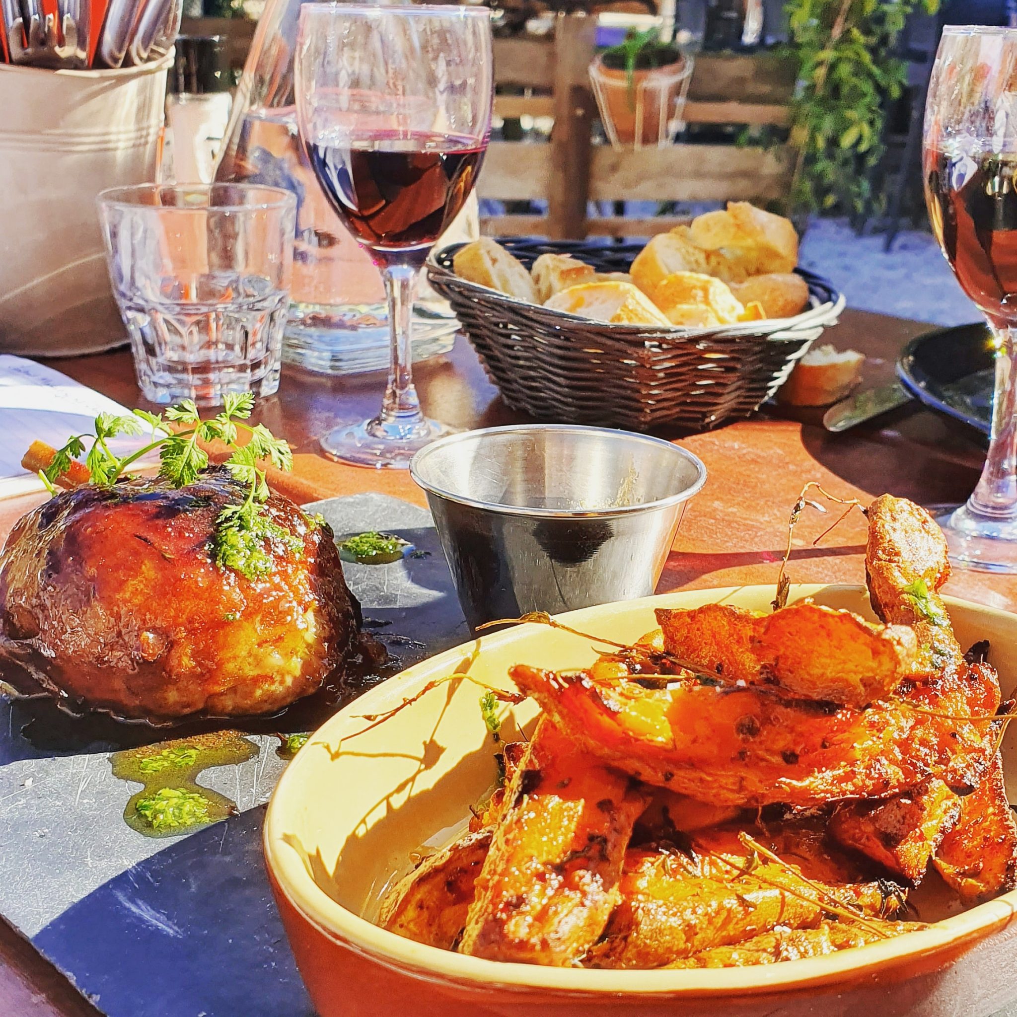 A foodie’s dream: a table set outdoors with roasted meat, seasoned potato wedges, red wine, bread, and water glasses—all brightly lit by sunlight. One of the best things to do in March 2020 for food lovers.