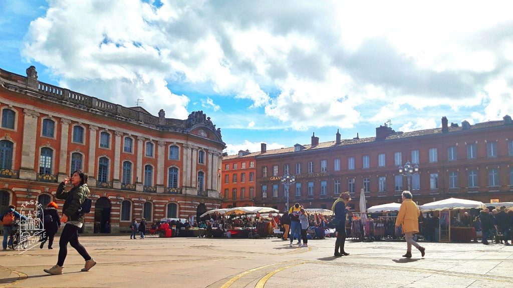 People walk across a sunlit plaza in Toulouse with red-brick buildings and market stalls under a partly cloudy blue sky. Some shop or stroll while others talk on the phone—perfect for Zoom virtual backgrounds that capture lively city life.