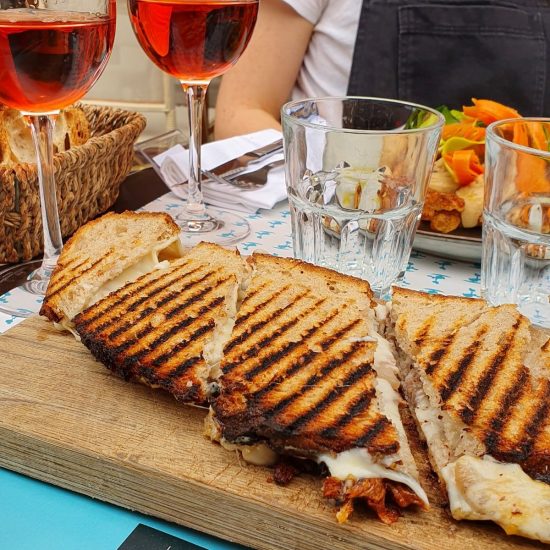 A grilled cheese sandwich cut into triangles sits on a wooden board. Two glasses of rosé wine, a salad, and a basket of bread are on the table—a true Foodie Things to Do experience in Toulouse. A person in a dark apron is partially visible in the background.