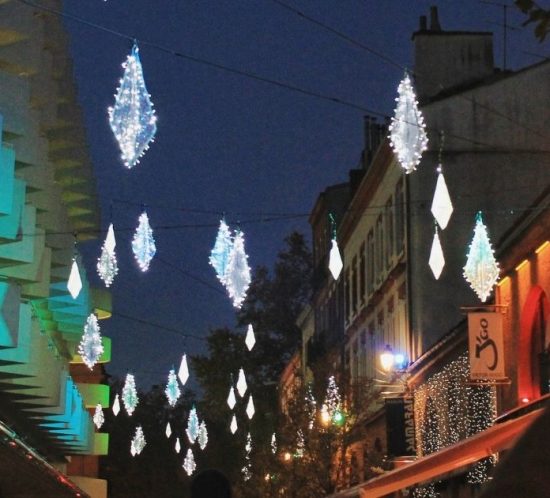 A street at night is decorated with glowing diamond-shaped lights overhead, creating a festive holidays vibe. People are taking photos, and buildings on both sides are illuminated with colorful lights, evoking the magic of virtual travel.