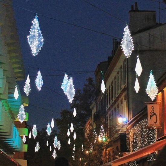 A street at night is decorated with glowing diamond-shaped lights overhead, creating a festive holidays vibe. People are taking photos, and buildings on both sides are illuminated with colorful lights, evoking the magic of virtual travel.