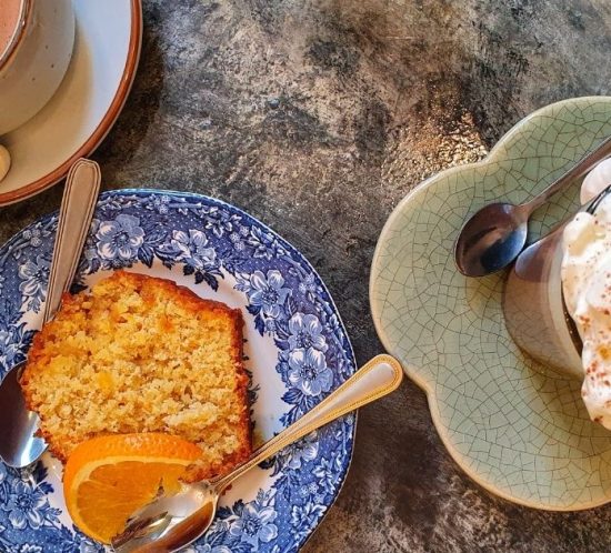 A blue floral plate with a slice of cake and an orange wedge, a cup of hot chocolate, and a glass topped with whipped cream and cocoa powder are set on a textured table—perfect foodie things to do during lockdown in November 2020.