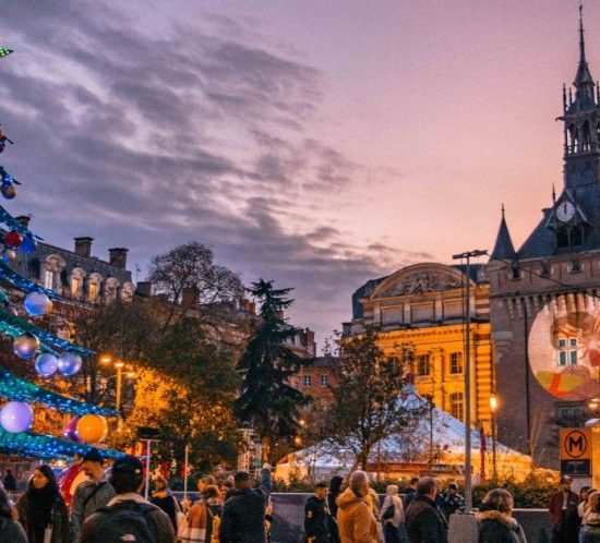 A decorated Christmas tree with a star topper stands in a lively square at dusk during Christmas in Toulouse, near a historic building with a tall clock tower and festive lights—perfect for free backgrounds.