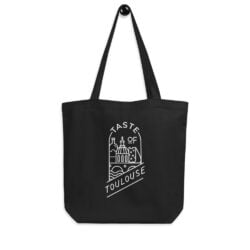 The Tote Bag with White Logo hangs on a wall hook, displaying a white logo and line drawing of Toulouse landmarks, with "Taste of Toulouse" in bold letters across the lower half.