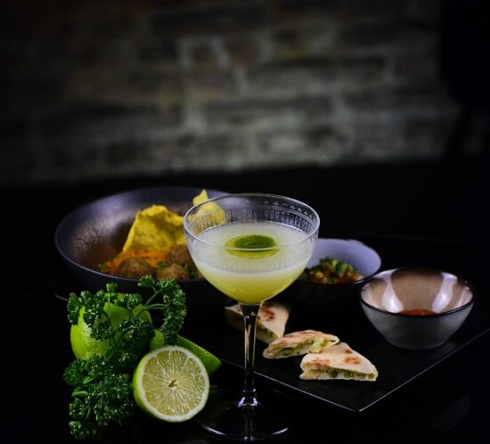 A cocktail glass with a lime slice sits on a table next to sliced lime, fresh herbs, naan bread, and bowls of sauces and curry—perfect for romantic dining inspired by a Toulouse dining guide, all set against a rustic stone background.