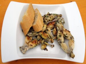 A white plate with grilled frog legs—a classic of French cuisine—garnished with herbs and served with two pieces of crusty baguette on an orange surface. One of the unique foods to try in Toulouse.
