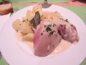 A plate of sliced ham covered in creamy white sauce, garnished with chopped herbs, served with boiled potatoes—a classic comfort dish you might find in Toulouse, France. A slice of bread and a fork are placed beside the plate.