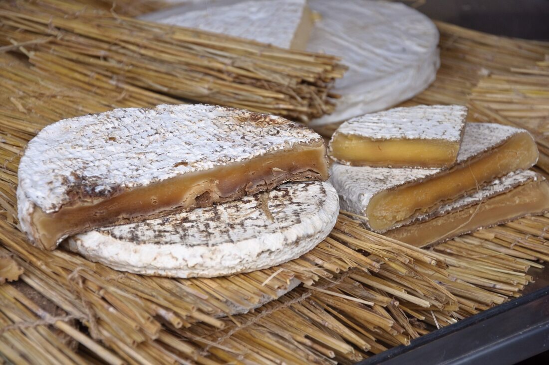 A round cheese with a white, moldy rind and a cut wedge revealing a soft, gooey brownish-yellow interior, displayed on dried straw—one of the strange foods France has to offer. Another round cheese and wedges are in the background.