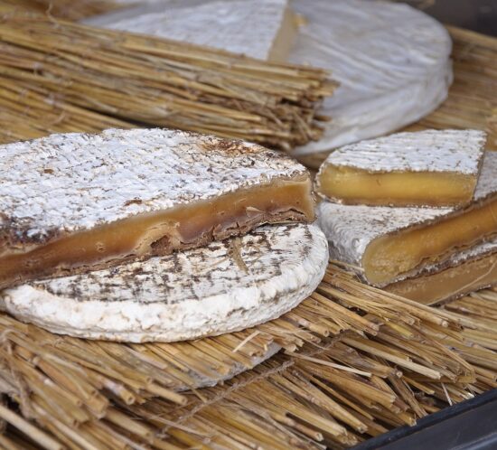 A round cheese with a white, moldy rind and a cut wedge revealing a soft, gooey brownish-yellow interior, displayed on dried straw—one of the strange foods France has to offer. Another round cheese and wedges are in the background.
