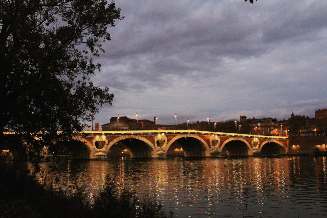 A stone bridge with arches in Toulouse is illuminated by lights at dusk, reflecting on a calm river. Trees frame the left, and city buildings are visible in the background under a cloudy sky—one of the top tourist attractions and things to do in the city.