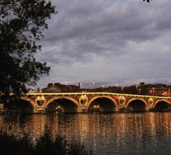 A stone bridge with arches in Toulouse is illuminated by lights at dusk, reflecting on a calm river. Trees frame the left, and city buildings are visible in the background under a cloudy sky—one of the top tourist attractions and things to do in the city.