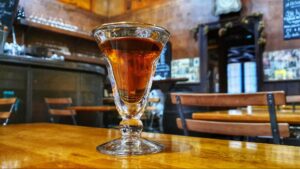 A close-up of a glass filled with a dark amber drink sits on a wooden table in a cozy, warmly lit café in Toulouse—an inviting stop among the city's top things to do and tourist attractions, with wooden chairs, tables, and a bar in the background.
