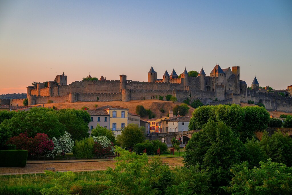 Carcassonne castle with stone walls and many towers on a hill at sunset, overlooking the town with colorful houses and lush green trees in the foreground—a perfect destination for an unforgettable day trip from Toulouse.