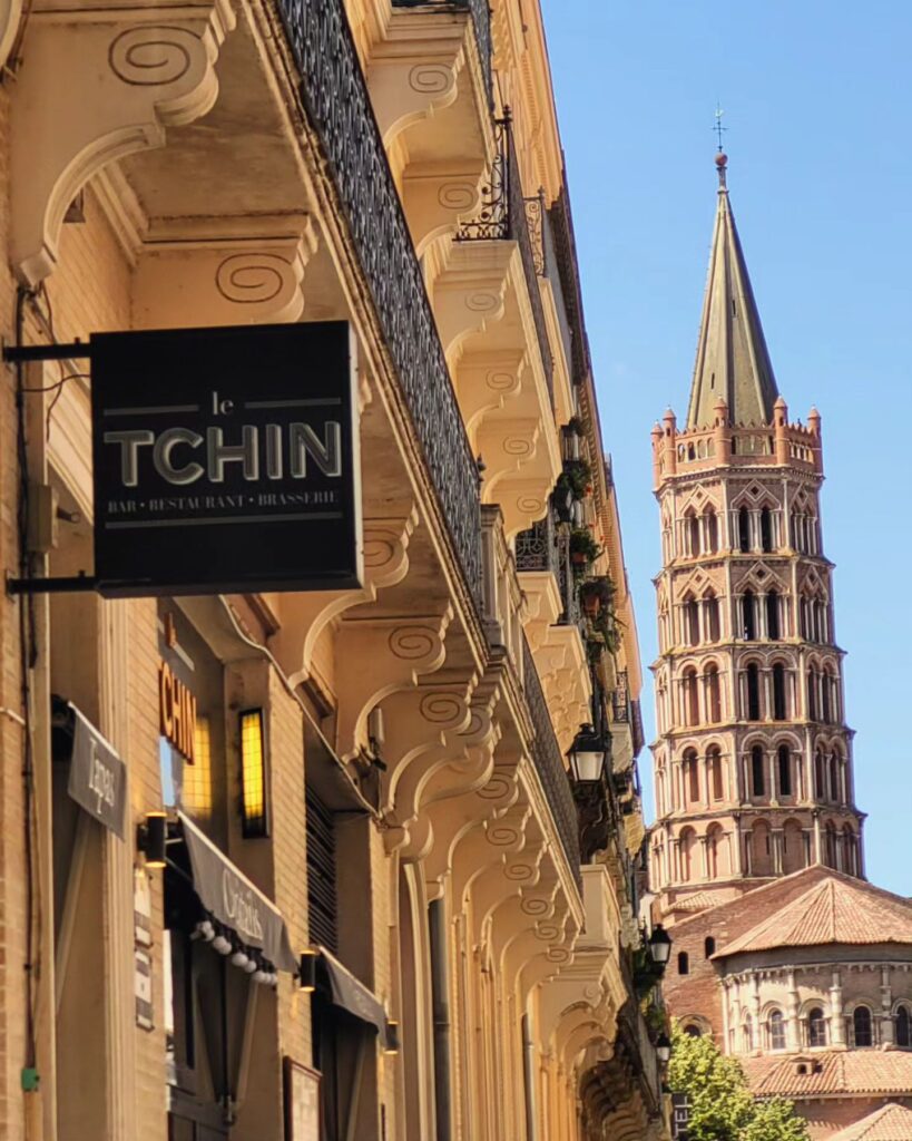 A tall, ornate church tower rises behind a row of yellow buildings with balconies and a black "Le Tchin" restaurant sign in the foreground—an inviting spot to eat dinner Toulouse-style before 7 pm under a clear blue sky.