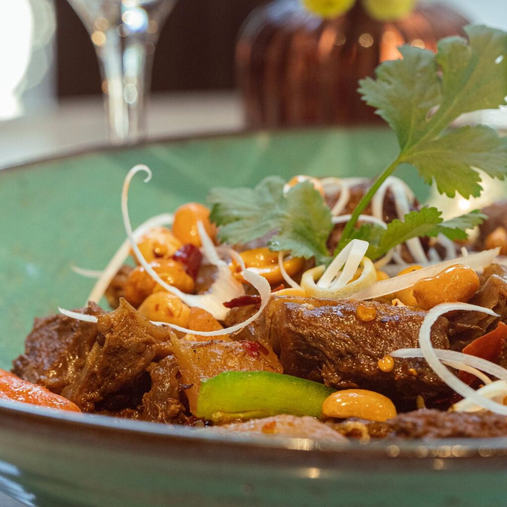 A close-up of a bowl filled with braised meat, peanuts, green bell peppers, carrots, bean sprouts, and cilantro. Perfect for those looking to Eat Dinner in Toulouse or explore popular Toulouse Restaurants. Served in a green bowl with a blurred background.