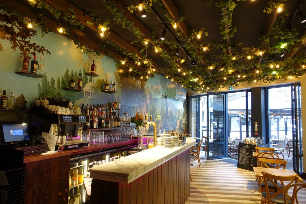 A cozy Toulouse restaurant bar with wooden furniture, a marble countertop, string lights, leafy ceiling decor, and a colorful nature mural. Sunlight streams through open glass doors to the patio—perfect for dinner before 7 pm.