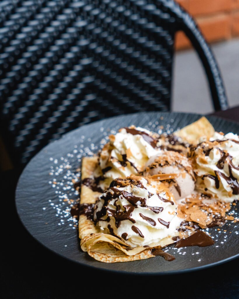 A black plate with a folded crepe topped with vanilla ice cream, whipped cream, chocolate sauce, and powdered sugar—perfect for anyone seeking early dinner in Toulouse, set on a table with a woven chair in the background.