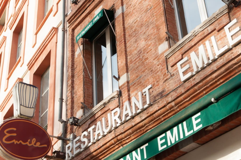 A close-up view of a brick building in Toulouse with white letters spelling "RESTAURANT EMILE" and a green awning. A round, red and gold sign with "Emile" hangs beside the entrance—perfect for those wondering where to eat cassoulet.