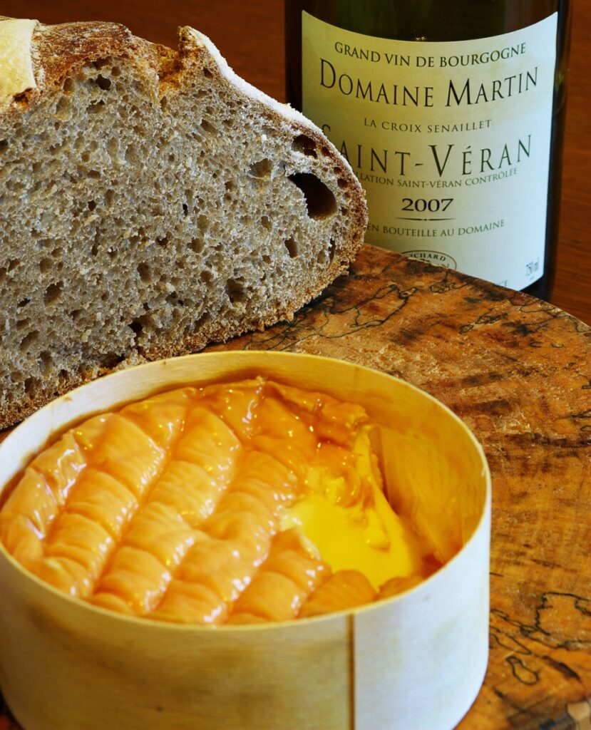 A round of creamy cheese in a wooden box sits on a wooden board next to a rustic loaf of bread and a bottle of Domaine Martin Saint-Véran, inviting the perfect cheese and wine pairing experience.