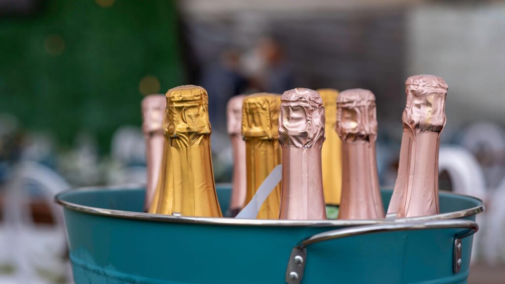 Bottles of champagne with gold and pink foil tops are chilled in a teal ice bucket, ready for celebration. The blurred background hints at an outdoor event, perfect for a festive cheese and wine pairing experience.