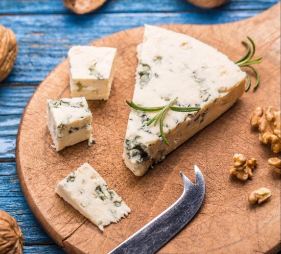 A wedge and cubes of blue cheese with rosemary on a round wooden board, surrounded by walnuts and a cheese knife, set on a rustic blue wooden table—perfect for an elegant cheese and wine pairing.