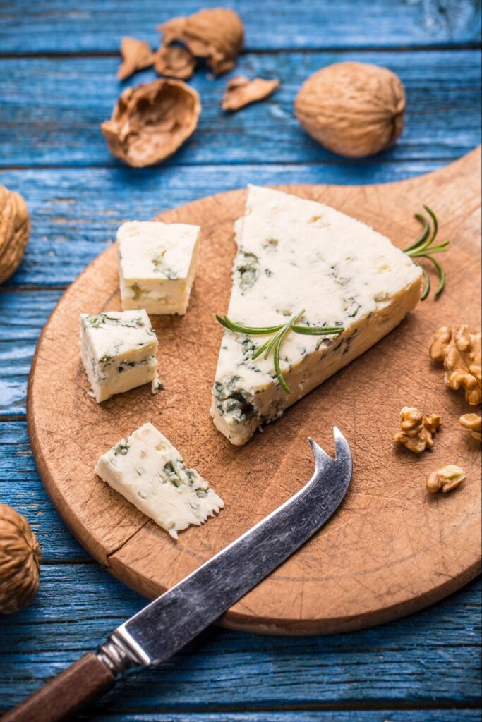 A wedge and cubes of blue cheese with rosemary on a round wooden board, surrounded by walnuts and a cheese knife, set on a rustic blue wooden table—perfect for an elegant cheese and wine pairing.