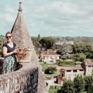 A woman wearing sunglasses and a patterned skirt stands on a stone balcony with a large basket, smiling towards the camera during an Albi Gourmet Food Tour. Behind her are pointed rooftops and a scenic view of trees and buildings in Albi.