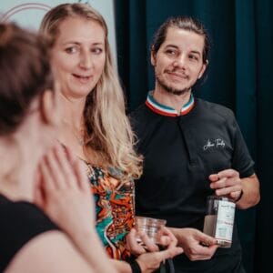 Three people are talking together indoors. One woman in a colorful dress is speaking about an Albi Gourmet Food experience, while another listens with her hand raised and a man in a dark shirt holds a glass, smiling at them.